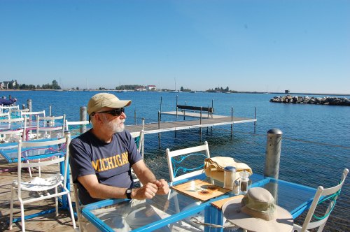 A man in a Michigan t-shirt sits at a table by the water, with a clear blue sky and boats in the background.