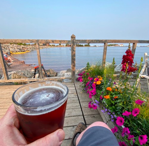 A person holds a beer while sitting on a deck overlooking a calm lake, surrounded by colorful flowers.