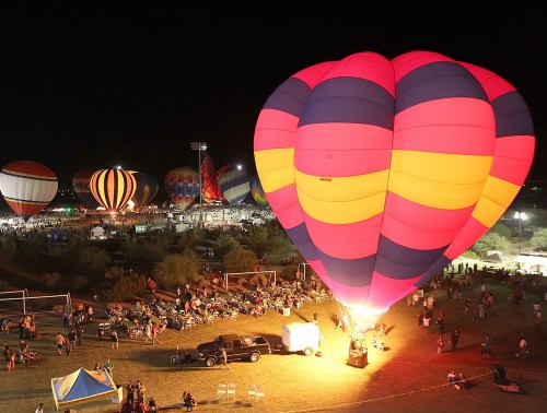 A brightly colored hot air balloon illuminates the night sky at a festival, surrounded by spectators and other balloons.