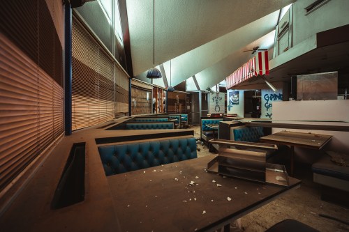Abandoned restaurant interior with empty booths, scattered debris, and graffiti on the walls. Natural light filters in.