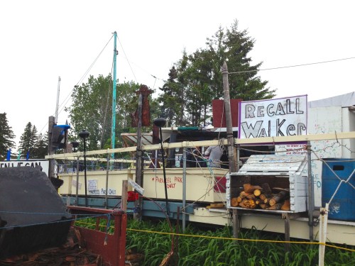 A makeshift structure with signs reading "Recall Walker," surrounded by trees and stacked firewood.
