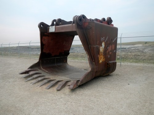 A large, rusty excavator bucket sits on a gravel path, with a fenced construction site in the background.