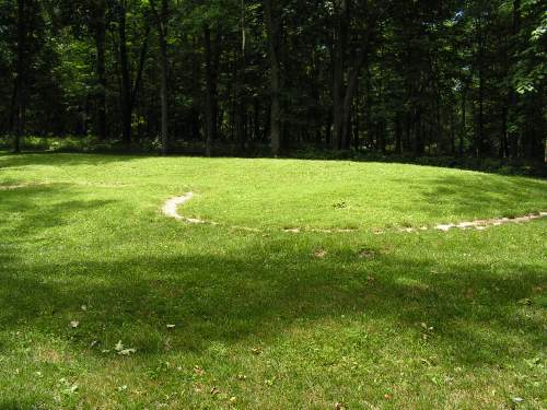 A grassy circular area in a forest, featuring a winding path and dappled sunlight filtering through the trees.