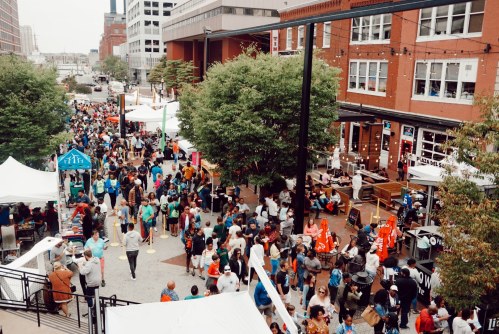 A bustling outdoor event with crowds, tents, and food stalls in an urban setting. Trees and buildings are visible.