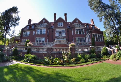 A large, elegant brick mansion with multiple gables, surrounded by lush gardens and greenery under a clear blue sky.