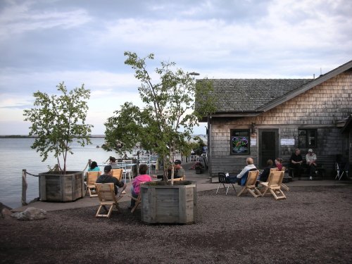A lakeside scene with people sitting in wooden chairs near a rustic building and trees, enjoying the view.
