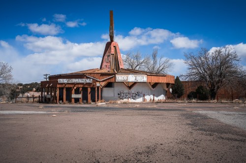 Abandoned Taco Bell building with a distinctive roof, surrounded by barren land and a blue sky with clouds.