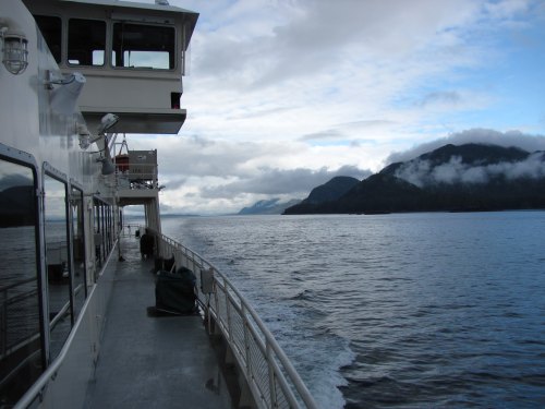 A ferry deck with a view of calm waters and mountains under a cloudy sky.