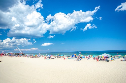 A sunny beach scene with colorful umbrellas, people relaxing on the sand, and a vibrant blue sky with clouds.
