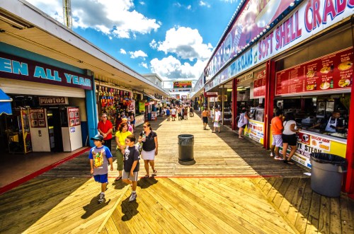 A bustling boardwalk scene with people walking, colorful stalls, and bright blue skies with fluffy clouds.