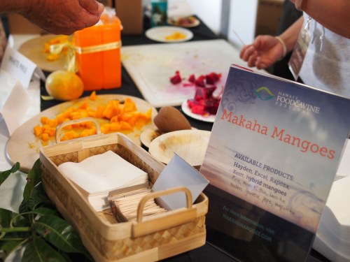 A table with cut mangoes and other fruits, featuring a sign for Makaha Mangoes and available products.