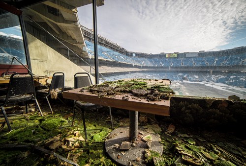 Abandoned stadium interior with a table covered in moss and debris, overlooking empty, dilapidated seating.