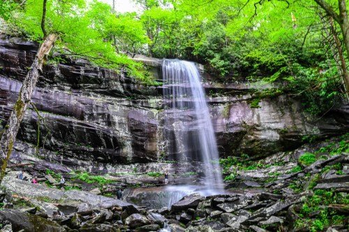 A man standing behind the 80-foot plunge fall of Rainbow Falls as it glistens in the sunlight surrounded by rocks and greenery of the forest.