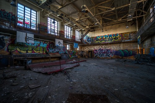 Abandoned gymnasium with colorful graffiti on walls, scattered debris, and sunlight streaming through large windows.