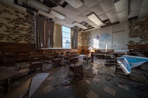 Abandoned classroom with overturned desks, debris on the floor, and sunlight streaming through a large window.