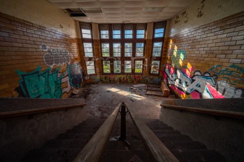 Abandoned interior with graffiti-covered walls, large windows, and a staircase leading down to a dusty floor.