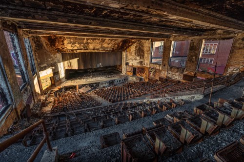 Abandoned theater interior with worn seats, a dusty stage, and peeling walls, illuminated by soft natural light.