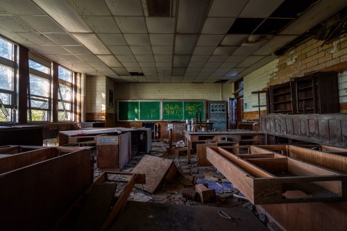 Abandoned classroom with scattered debris, overturned furniture, and graffiti on the chalkboard. Sunlight filters through windows.