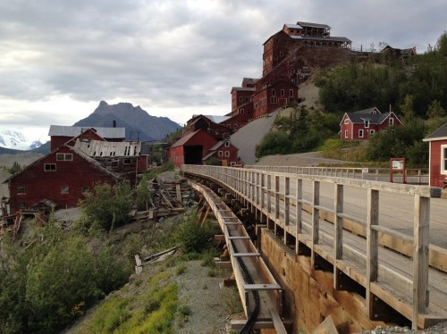 Abandoned red buildings on a hillside with a wooden walkway, surrounded by greenery and mountains under a cloudy sky.
