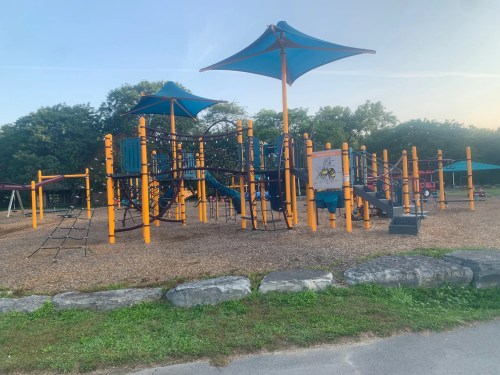 Colorful playground with climbing structures and shaded areas, surrounded by gravel and greenery.