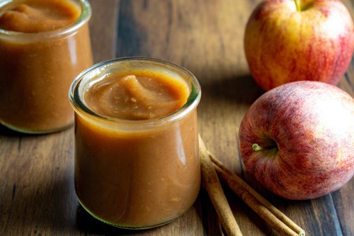 Two jars of apple butter beside fresh apples and cinnamon sticks on a wooden surface.