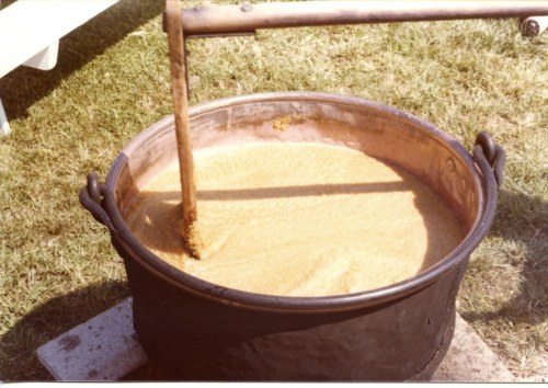 A large pot filled with a thick, golden mixture, with a stirring stick resting on the edge. Grass is visible in the background.