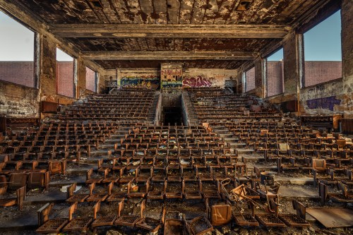 Abandoned auditorium with rusted chairs, graffiti on walls, and debris scattered across the floor.