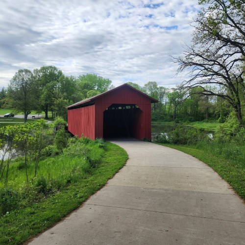 A red covered bridge stands at the end of a paved path, surrounded by lush greenery and a calm pond.