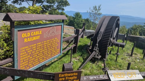Sign for the Old Cable Railway with a large gear, surrounded by greenery and mountains in the background.