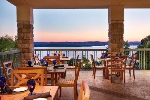 A scenic restaurant patio with wooden tables and blue glassware, overlooking a tranquil lake at sunset.