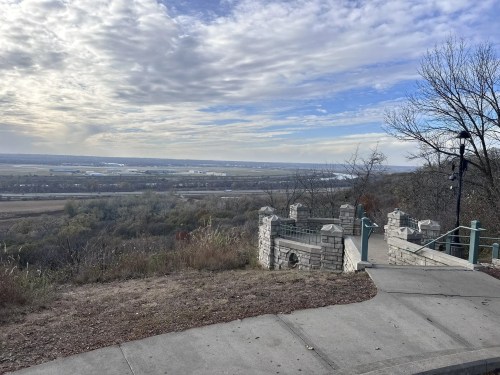 A scenic overlook with a stone railing, featuring a view of a river valley and cloudy sky in the background.