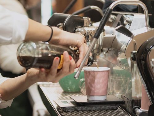 A barista pours syrup into a pink cup while using an espresso machine, with a scale visible on the counter.