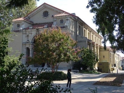 A historic building with a decorative facade, surrounded by trees and a pathway, with a person walking nearby.
