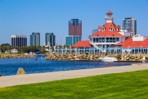 A waterfront view featuring a red-roofed building, boats, and a skyline of modern skyscrapers under a clear blue sky.