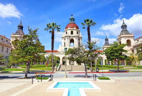 A grand building with a dome and palm trees, featuring a fountain in the foreground and a blue sky above.