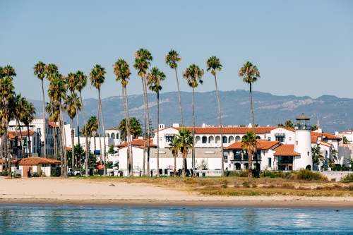 A coastal scene featuring palm trees, white buildings, and a lighthouse against a backdrop of mountains and clear blue sky.