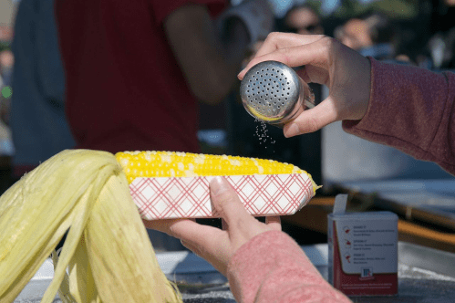 Person shaking salt on an ear of roasted corn that they are holding.