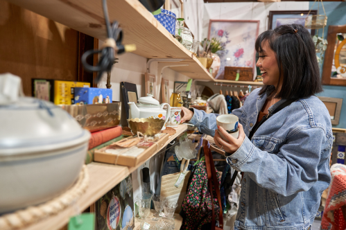A woman in a denim jacket examines vintage items on a wooden shelf in an antique shop.