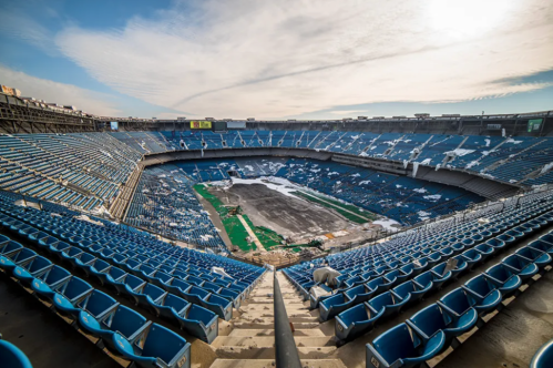 An empty stadium with blue seats, a covered field, and scattered snow under a cloudy sky.