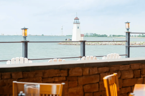 A lighthouse stands by the water, with a view of a rocky shore and chairs in the foreground. Overcast sky.