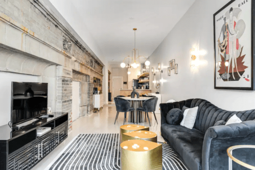 Modern living room featuring a black sofa, gold accent tables, a TV, and stylish decor with a dining area in the background.