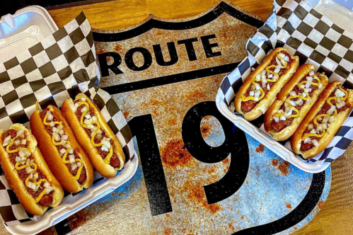 Two trays of hot dogs with mustard and onions on a rustic Route 19 sign background.