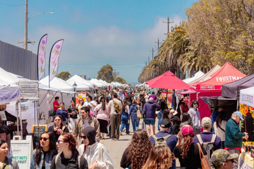 A bustling outdoor market with colorful tents, people browsing, and palm trees under a clear blue sky.