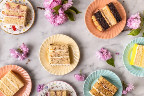 A variety of cake slices on colorful plates, surrounded by pink flowers on a marble surface.