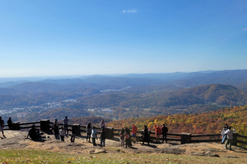 A scenic overlook with people enjoying a vibrant autumn landscape of mountains and valleys under a clear blue sky.