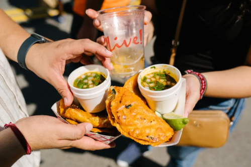 Two hands hold a plate with golden empanadas and cups of soup, alongside a drink in a clear cup.