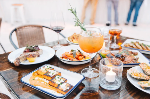 A table set with various dishes, including grilled meats, sides, and a colorful cocktail garnished with a sprig of rosemary.