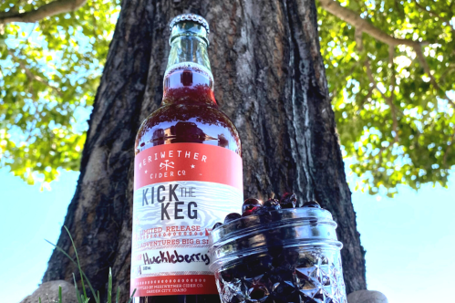 A bottle of Kick the Keg huckleberry cider next to a jar of huckleberries, with a tree in the background.