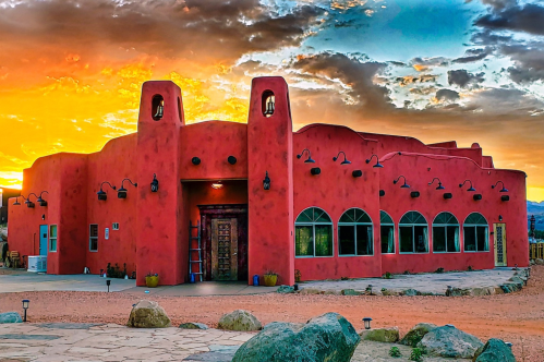 A vibrant red adobe building with arched windows, set against a colorful sunset sky and rocky landscape.