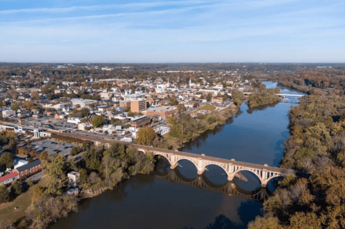 Aerial view of a river winding through a town, featuring a stone bridge and autumn-colored trees along the banks.